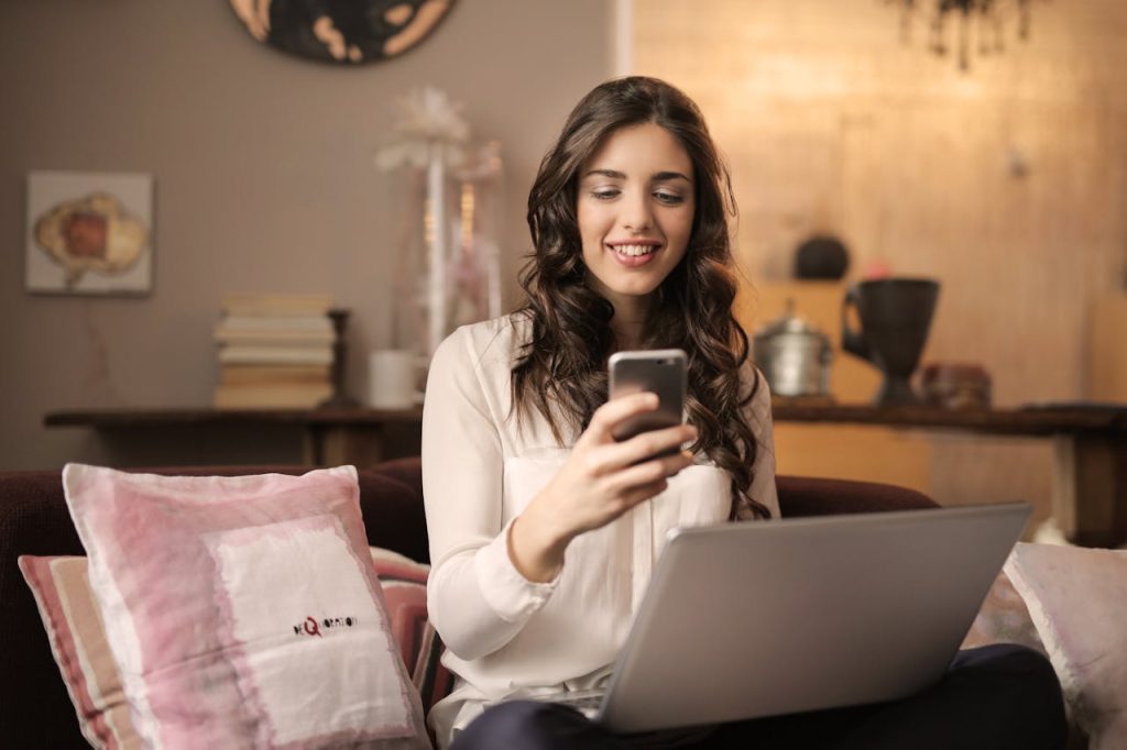 woman-sitting-on-sofa-while-looking-at-phone-with-laptop-on-lap-920382 A woman enjoying leisure time using her smartphone and laptop in a cozy living room.